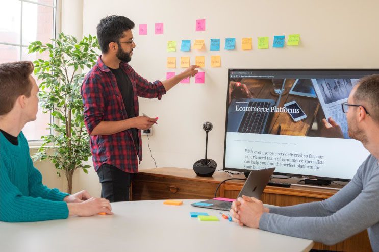 Two male colleagues are sat at a table watching a male colleague pointing to post it notes on a wall.