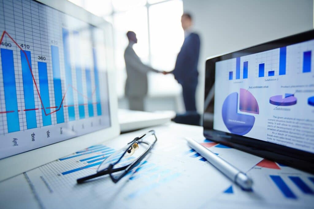 Desk with glasses, pen and data analysis charts printed on paper also displayed on tablet screen. Two males are shaking hands in the background.