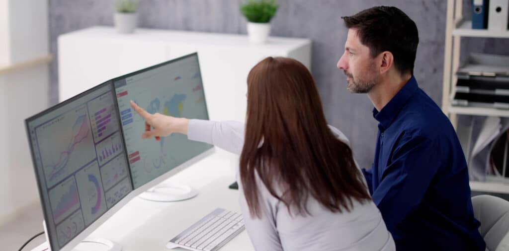 A female and male sat beside each other at a desk. The female is pointing to business data on a screen and male is looking at this.