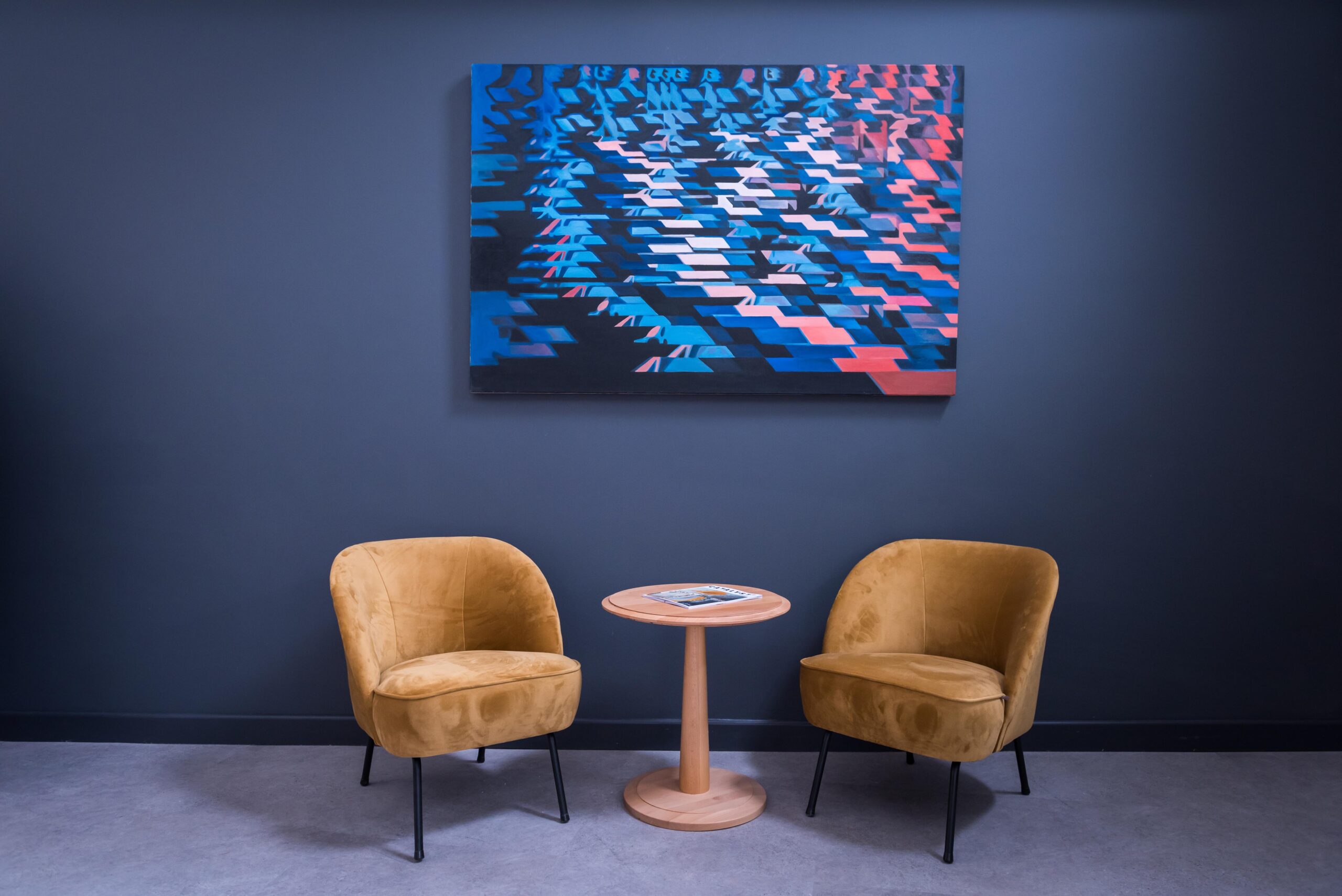 Two mustard chairs with a table in the middle of them. Art work is displayed on a grey wall above the table and chairs.